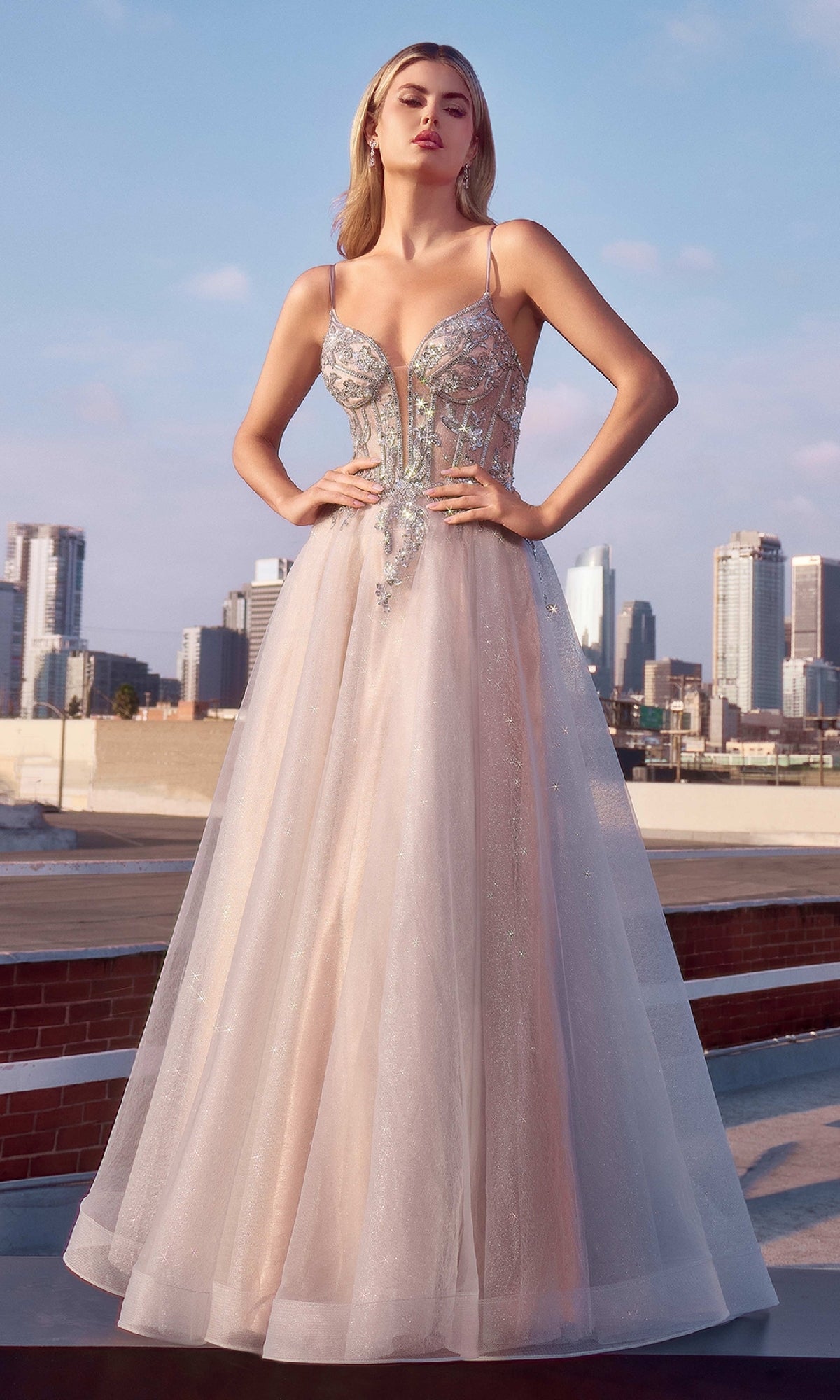 A woman in the Ladivine CC0881 Sheer-Corset Long Prom Dress, featuring sparkling beaded embroidery and thin straps in light pink, stands on a rooftop with a city skyline in the background.